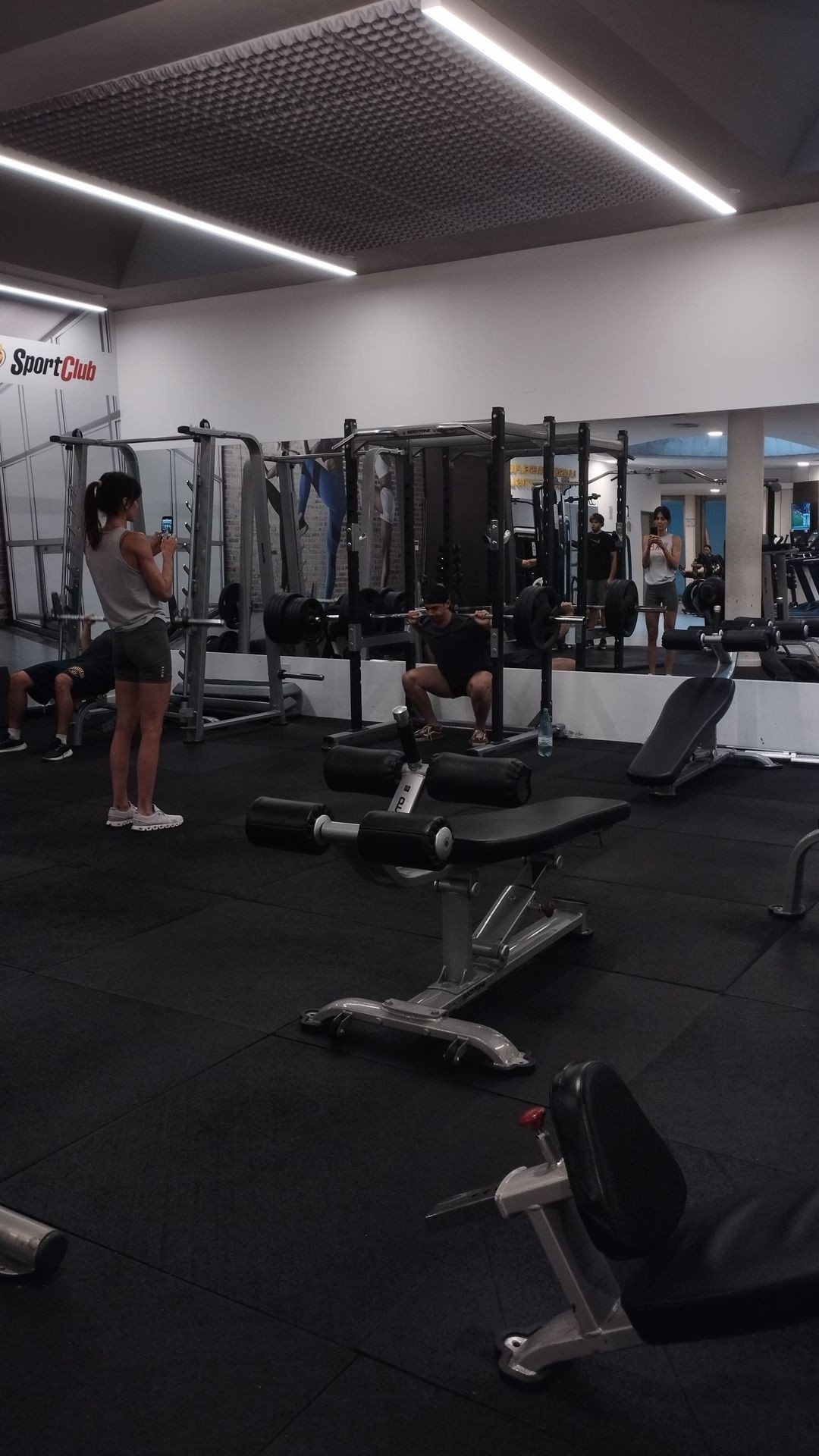 People working out with various fitness equipment in a gym, including weight benches and a squat rack.